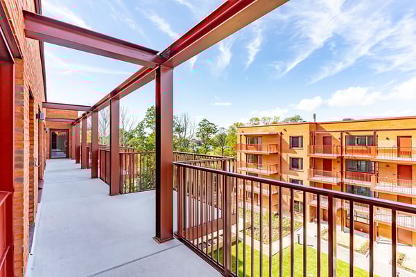 Utsikt över en modern balkong med röda metallräcken och byggnader av tegel samt grönska i bakgrunden under en klar blå himmel