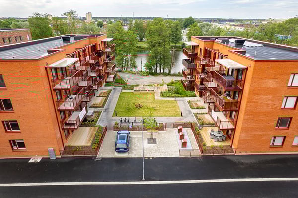 Lägenhetskomplex med två orangea byggnader, innergård med gräsmatta och planteringar, parkeringsplats med en blå bil, och en å i bakgrunden