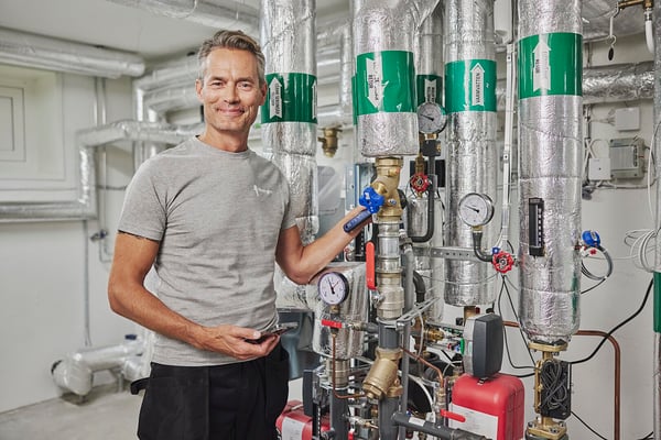 Man i grå t-shirt står i ett tekniskt rum med rör och tryckmätare, omgiven av isolerade rör och utrustning