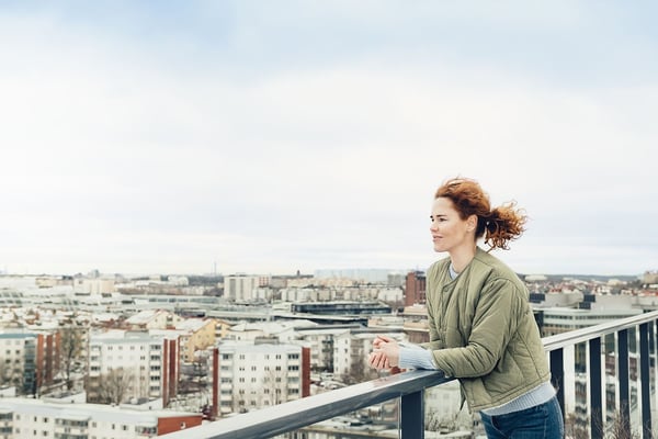 Person som lutar sig mot ett räcke på en balkong med utsikt över en stad i bakgrunden, en kylig vinterdag med snö på taken