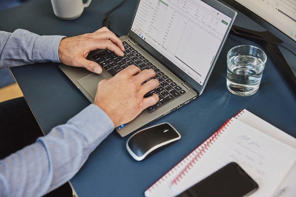 Person skriver på en bärbar dator med ett kalkylblad öppet, omgiven av en anteckningsbok, smartphone, mus och ett glas vatten på ett mörkblått bord