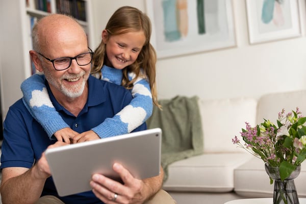 En man i blå pikétröja håller i en surfplatta medan ett barn i en blå- och vitrandig tröja står bakom honom i ett vardagsrum med ljusa möbler och konst på väggarna