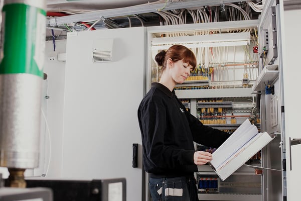 Person arbetar vid ett öppet elskåp och håller i en pärm, omgiven av kablar och tekniska komponenter