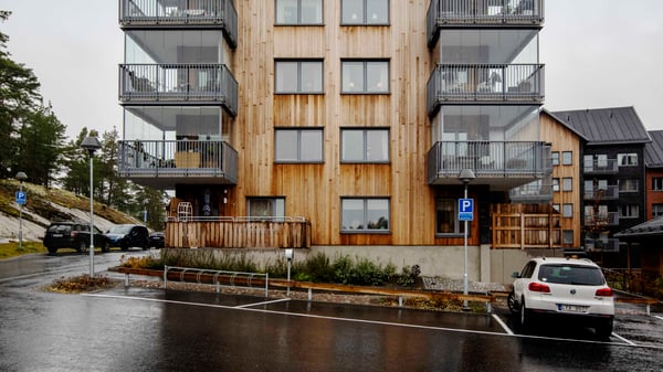 Flerfamiljshus med träfasad och balkonger, omgiven av parkeringsplatser och grönska på en regnig dag