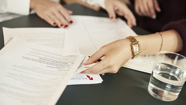 Person räcker fram dokument på ett bord med ett glas vatten och fler papper i bakgrunden