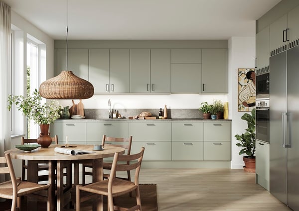 Kök med ljusa grönskande skåp, rostfria vitvaror, en rund matbord med trästolar och en hängande rottinglampa, omgiven av växter och naturligt ljus från ett stort fönster