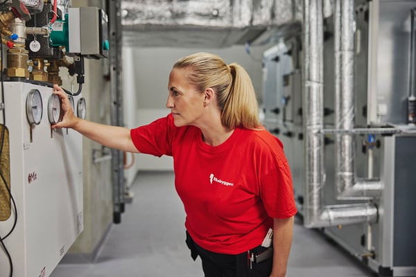 En person i röd t-shirt står i en teknisk anläggning och kontrollerar en mätare på en maskin, omgiven av rör och maskinell utrustning