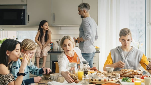 En grupp människor runt ett matbord i ett modernt kök, med tallrikar, glas, frukostmat och drycker synliga på bordet. En person står vid diskbänken i bakgrunden