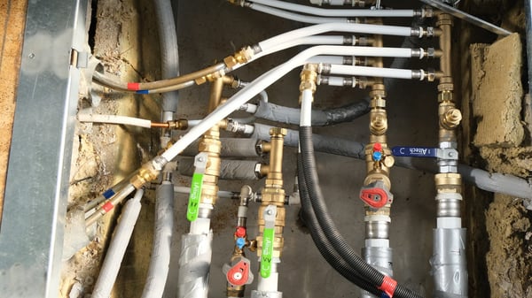 Rörinstallation med flera kopplingar, ventiler och anslutningar, inklusive vita och grå slangar, monterade på en vägg med delvis synlig isolering