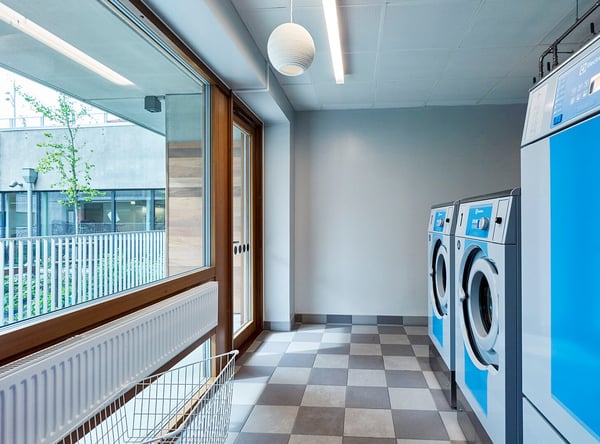 Tvättstuga med blå tvättmaskiner, rutigt golv och stora fönster som släpper in naturligt ljus