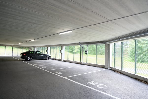 Parkeringsgarage med en mörk bil parkerad på en av de markerade platserna, omgiven av glasväggar som visar grönska utanför