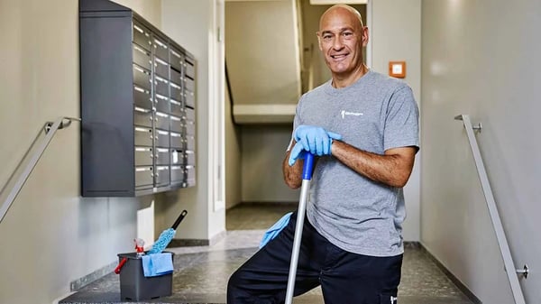 En person i arbetskläder håller i ett moppskaft i en trappuppgång med postboxar och städutrustning i bakgrunden