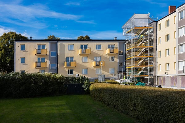 Flerbostadshus med gula balkonger och fasadrenovering på en del av byggnaden. Gräsmatta och häck i förgrunden under en klarblå himmel