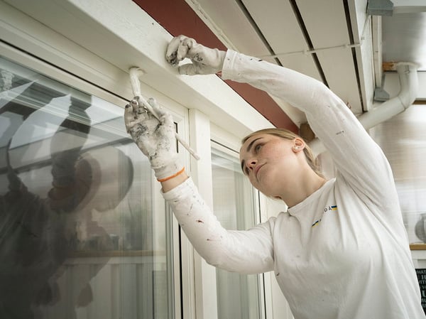 En person målar en träkarm runt ett fönster med en pensel; händer och kläder är täckta av vit färg