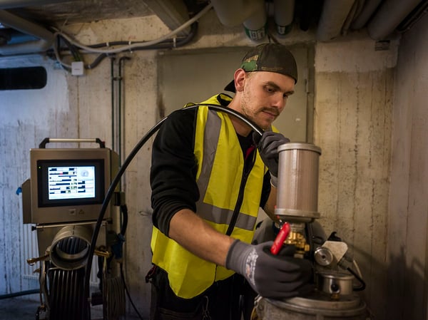 Person i gul reflexväst och gråa handskar som arbetar med en maskin i en industriell miljö, omgiven av rör och teknisk utrustning
