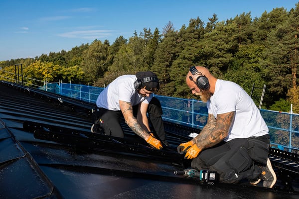 Två personer arbetar på ett tak med taksäkerhetsutrustning, omgivna av träd och skog i bakgrunden