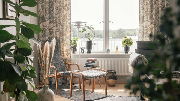 Vardagsrum med fåtölj, fotpall, växter och stora fönster med ljusa gardiner