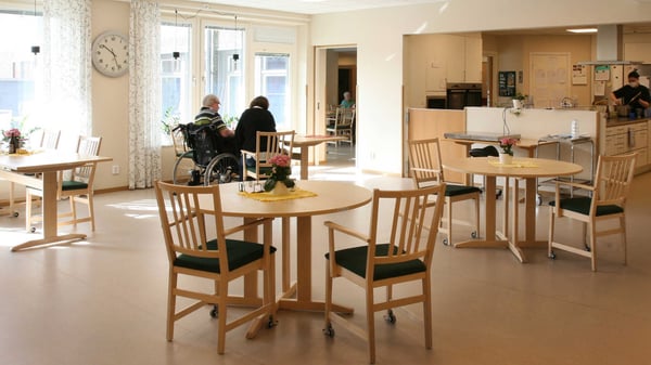 Gemensam matsal med runda bord, trästolar och några personer som sitter och samtalar