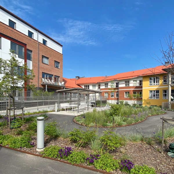 Bostadsgård med flerbostadshus, grönskande planteringar och röda tak under blå himmel