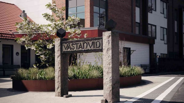 Skylt med texten Västanvid framför bostadshus och blommande trädgårdsgata