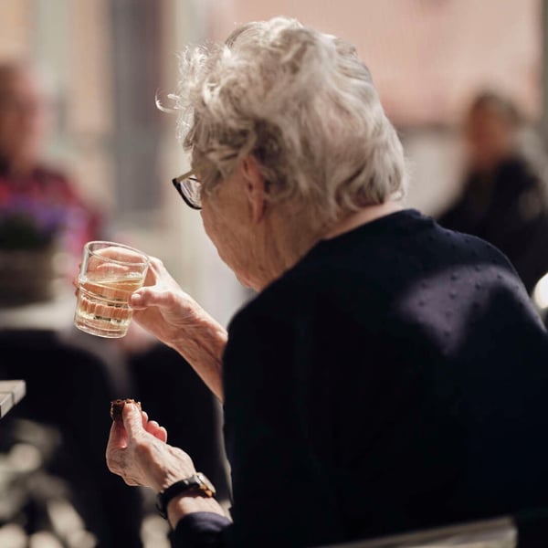 Äldre kvinna med grått hår som håller ett glas vatten och ett litet bakverk