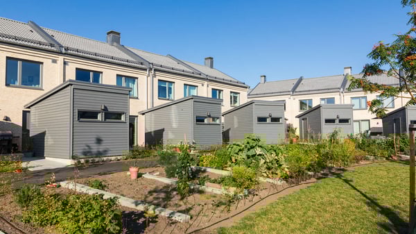 Inflyttningsklar lägenhet med små grå förrådsbyggnader och gemensam trädgård utanför flerfamiljshus