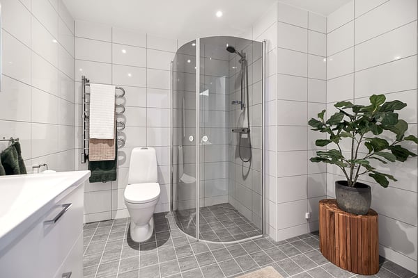 Modernt badrum med vita kakelväggar, duschhörna, toalett och grått klinkergolv
