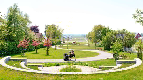Grön park med gångväg, träd och en person som promenerar med barnvagn