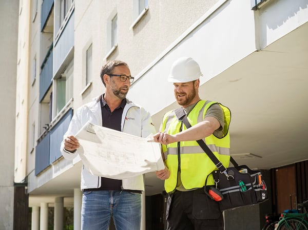 Två män står utomhus vid ett bostadshus och granskar en ritning. Den ena bär en gul varselväst och den andra en vit jacka