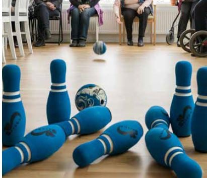 Klotträn med blå bowlingkäglor och en boll på ett golv, med personer sittande i bakgrunden