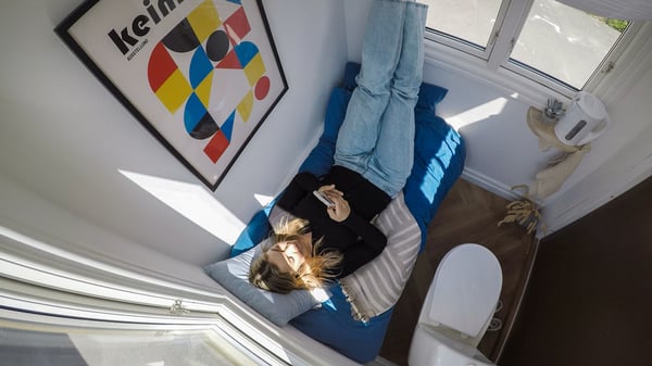 Person som ligger i en säng med blå madrass och kuddar, med telefon i handen och dagsljus från fönstret i ett minihus