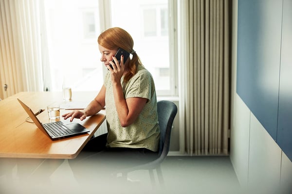 Kvinna vid ett bord som pratar i telefon och arbetar på en laptop i ett ljust rum