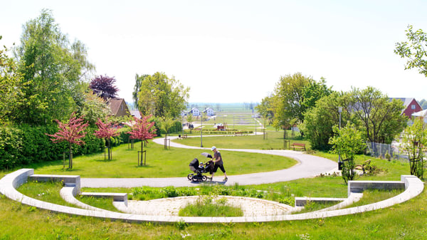 Grön park med gångväg, träd och en person som promenerar med barnvagn