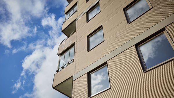 Fasaden på ett modernt flerbostadshus med stora fönster och balkonger mot en blå himmel med moln