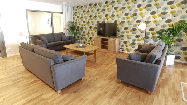 Gemensamt utrymme i äldreboende med grå soffor, soffbord, tv-bänk och mönstrad fondvägg