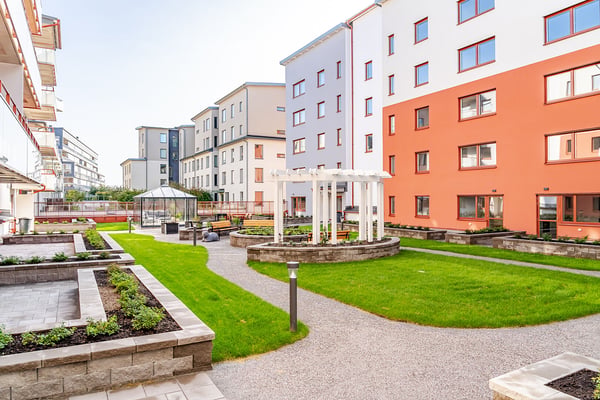 Modern bostadsområde med flerfamiljshus, grönskande gräsmattor, gångvägar och en pergola i centrum