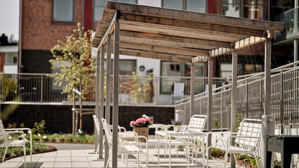 Tomt uteplatsområde med vita sittmöbler under pergola vid flerfamiljshus, inflyttningsklart