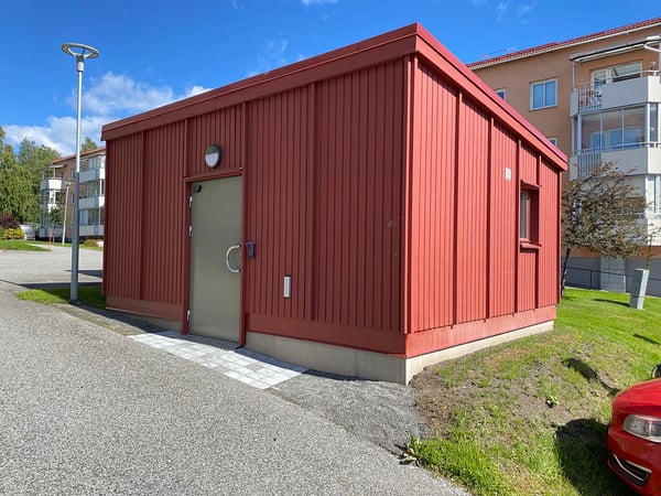 Rött miljöhus med dörr och fönster vid flerfamiljshus, omgivet av gångväg och gräsyta