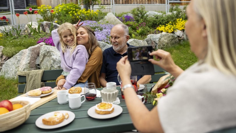En grupp människor sitter vid ett trädgårdsbord med tallrikar, koppar och bakverk, medan en person tar ett foto med en mobiltelefon. I bakgrunden syns blommor och stenar