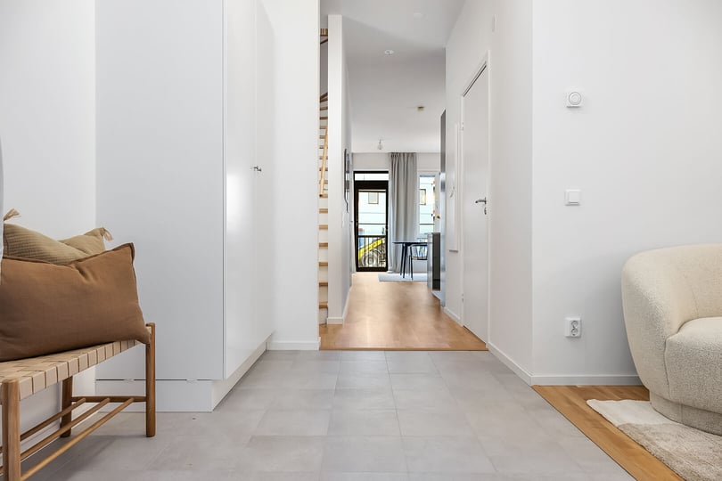 Hall med ljusa vita väggar, klinkergolv, en träbänk med kuddar till vänster och en beige fåtölj till höger. Utsikt mot en balkong genom glasdörrar i bakgrunden