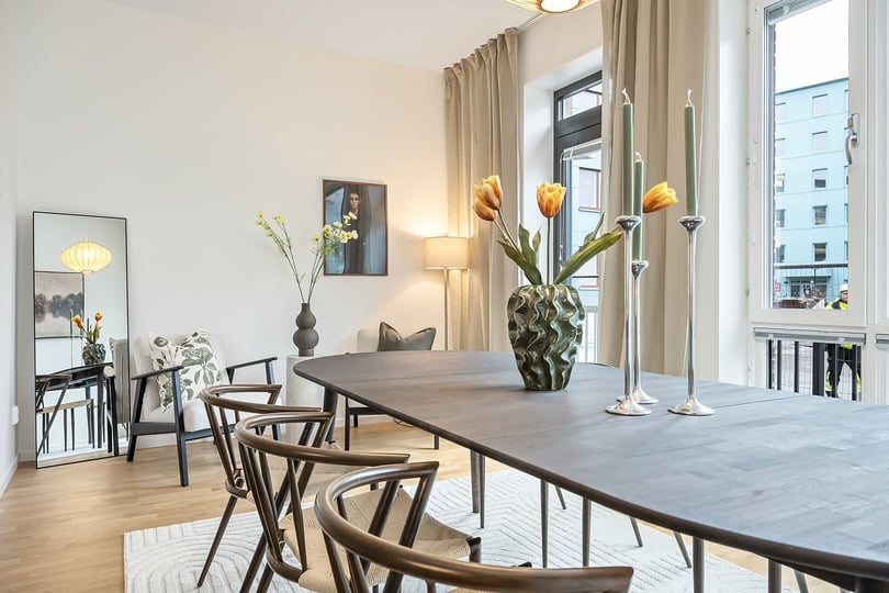 Matplats med mörkt träbord, designstolar och dekorationer som vas med gula tulpaner och ljusstakar. Bakgrund med stora fönster, spegel, väggkonst och gardiner i neutral färg