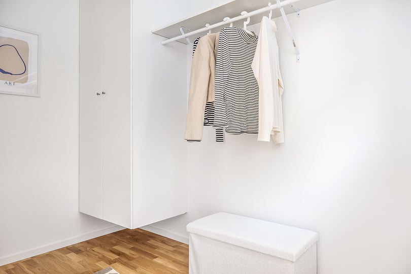 Vit garderob med hängande kläder, inklusive randig tröja och ljusa plagg, på en klädstång. Trägolv och minimalistisk inredning med en vit förvaringsbänk i förgrunden