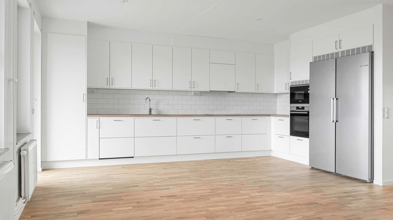 Vit modern köksinteriör med skåp, induktionsugn, inbyggd mikrovågsugn, kylskåp och diskbänk, samt trägolv och vita kakelplattor på väggen