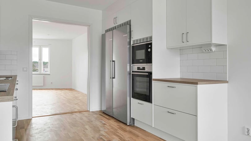 Modern kök med vita skåpluckor, rostfria vitvaror, mikrovågsugn, ugn och kylskåp. Öppet utrymme med trägolv leder till ett ljusfyllt rum i bakgrunden