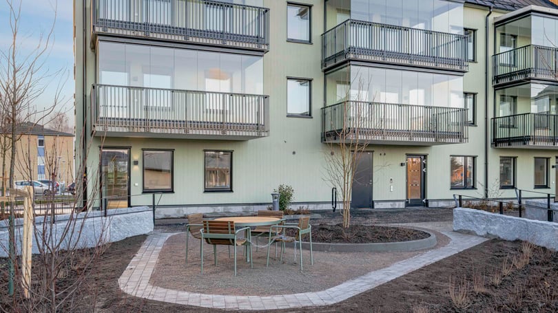 Flerbostadshus med gröna fasader, glasbalkonger och en liten uteplats med möbler omgiven av nyplanerade träd och buskar