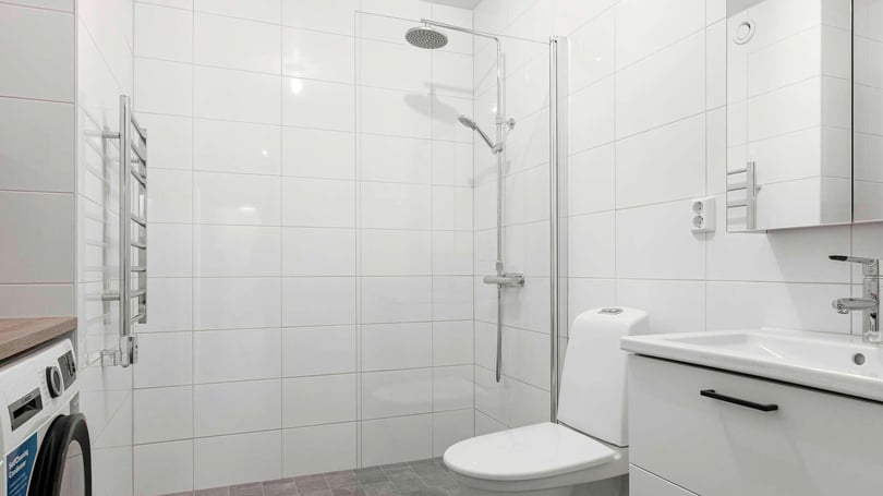 Badrum med vitkaklade väggar, dusch med glasvägg, toalett, tvättmaskin och vit handfat med spegelskåp