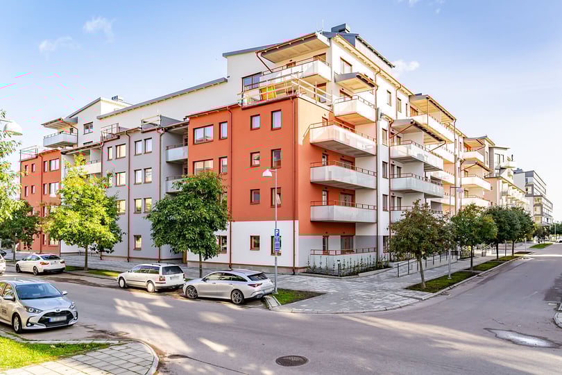 Flerfamiljshus med moderna fasader i vitt och orange, balkonger och parkerade bilar längs en gata kantad av träd