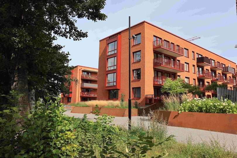 Moderna flerfamiljshus i tegel med röda balkonger, omgivna av grönska och gångvägar