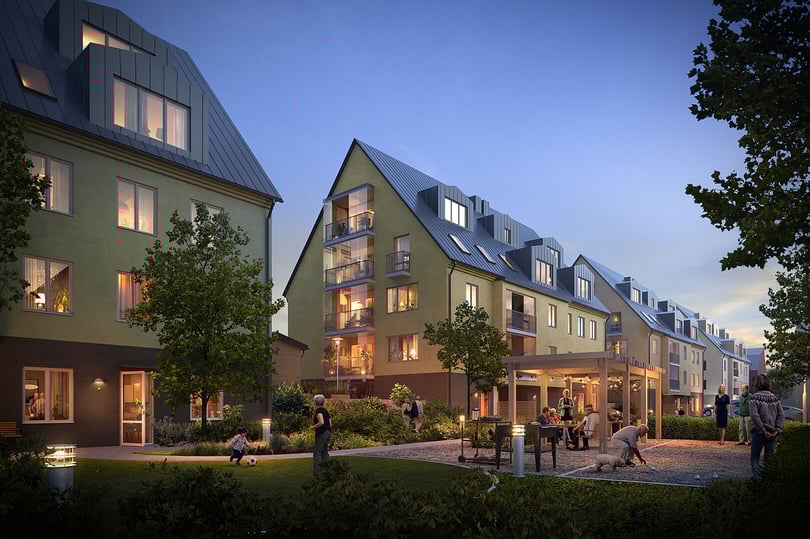 Modern bostadskvarter med gröna fasader, balkonger och en innergård med träd, gräsmatta och sittplatser i skymningsljus