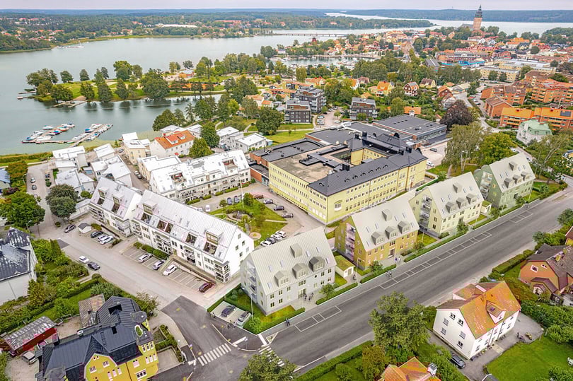 Flygbild över en stad vid en sjö med bostadshus, en skolbyggnad, grönområden och en småbåtshamn i bakgrunden